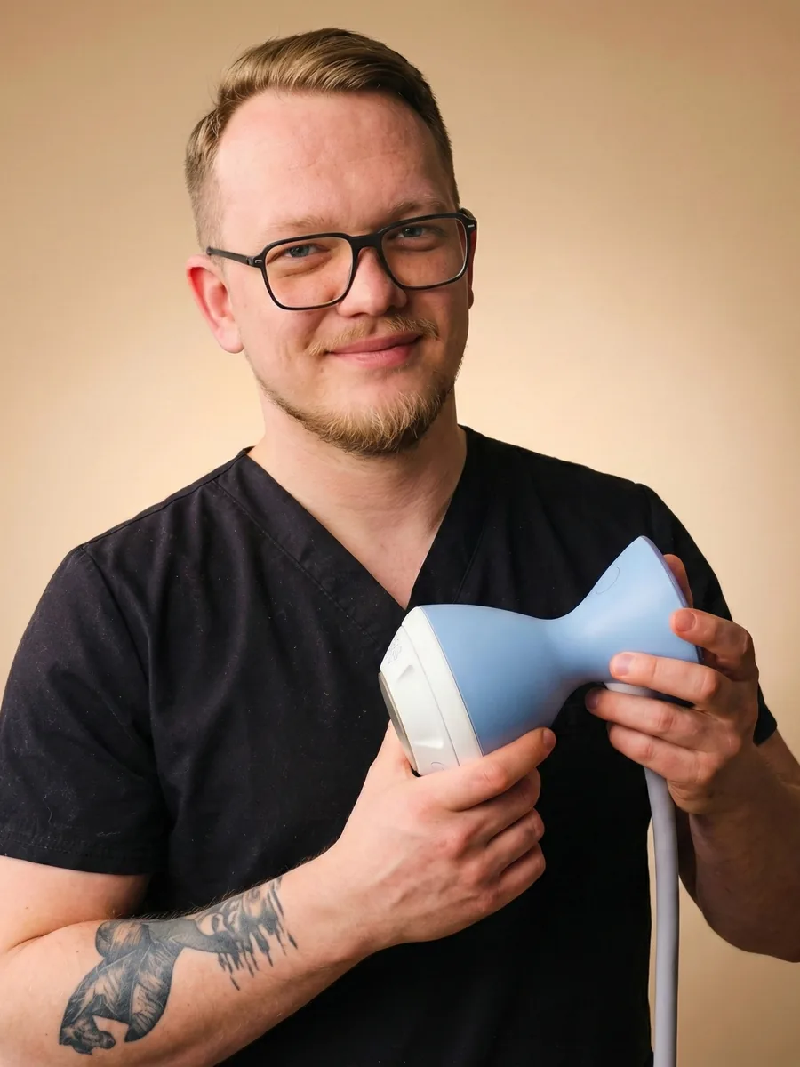 Fizjoterapeuta Marcin Jędrzejewski uśmiecha się, trzymając głowicę aparatu do fali uderzeniowej